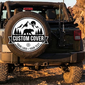 May include: A black and white spare tire cover with a mountain, bear, and tree design. The cover is on the back of a green Jeep. The text on the cover reads "CUSTOM COVER" and "YOUR LOGO - YOUR TEXT".
