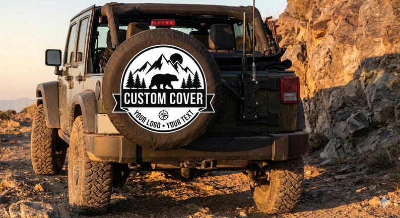 May include: A black and white spare tire cover with a mountain, bear, and tree design. The cover is on the back of a green Jeep. The text on the cover reads "CUSTOM COVER" and "YOUR LOGO - YOUR TEXT".