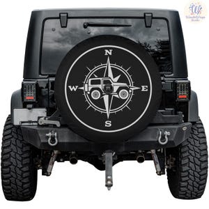 May include: Black Jeep with a spare tire cover featuring a white compass rose design and a Jeep silhouette. The compass points North, South, East, and West. The tire cover is mounted on the back of the black Jeep.