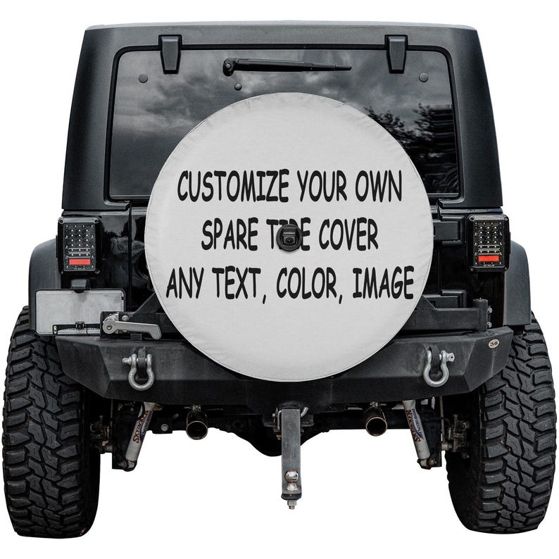 May include: A black Jeep SUV with a white spare tire cover. The cover features the text "CUSTOMIZE YOUR OWN SPARE TIRE COVER ANY TEXT, COLOR, IMAGE" in black. The vehicle has large, black tires and a black bumper.