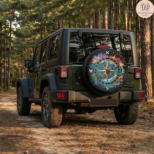 May include: A dark green off-road vehicle featuring a spare tire cover on the rear. The cover displays a colorful, artistic compass design with vibrant splashes of paint, mountain silhouettes, and the cardinal directions N, S, E, and W.