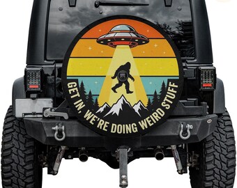 May include: Black Jeep with a spare wheel cover. The cover has a retro design of a UFO beaming up a Bigfoot, set against a sunset backdrop. Text reads "GET IN, WE'RE DOING WEIRD STUFF".