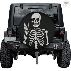 May include: Black Jeep with a spare tire cover featuring a white skeleton design. The skeleton is giving the rock and roll hand gesture. The Jeep has black tires and a black bumper.