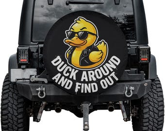 May include: Black spare wheel cover with a yellow cartoon duck wearing sunglasses and a black leather jacket. The text "DUCK AROUND AND FIND OUT" is printed in white. The cover is mounted on a black Jeep.