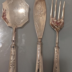 May include: A set of three silver-plated serving utensils with ornate handles. The set includes a serving spoon, a butter knife, and a fork.