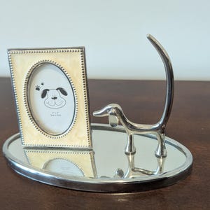 May include: A silver picture frame with a white and gold border and a dog silhouette inside. The frame is sitting on a silver tray with a silver dog sculpture.