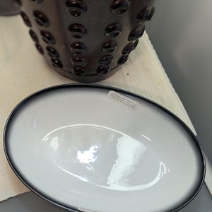 Peut inclure: Un plat de service ovale blanc avec un bord noir. Le plat présente un léger dégradé du blanc au noir autour du bord. Le plat est posé sur une surface blanche.