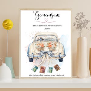 May include: A framed print featuring a vintage car decorated with flowers, a large bow, and money. The German text reads "Gemeinsam ist das schönste Abenteuer des Lebens" and "Herzlichen Glückwunsch zur Hochzeit!" The artwork has a watercolor style.