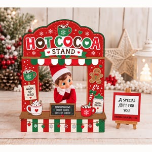 Puede incluir: Un letrero de madera rojo y verde de un puesto de chocolate caliente con una pequeña figura de elfo en el centro. El letrero muestra el texto HOT COCOA STAND, WARM UP HERE, MARSHMALLOWS CANDY CANES LOTS OF CHEER y un cartel A SPECIAL GIFT FOR YOU.