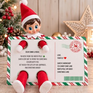 Pode incluir: Um boneco elfo de pelúcia com gorro e traje vermelho segura um cartão postal retangular com bordas de bengala doce vermelha e verde. O texto diz: Dear Emma & Noah, I'm here from the North Pole! Santa sent me to spend the Christmas season with you.