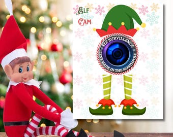 May include: A Christmas-themed image featuring an elf doll and a decorative sign. The sign reads "Elf Cam" and "Elf Surveillance" with a camera lens graphic. The elf doll is red and white, with black shoes. The sign is 5x7 inches.