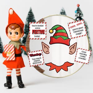 May include: A Christmas elf doll in a red outfit holding a gift. A decorative mirror shows an elf hat, ears, and collar, with text that reads "You're lookin' extra FESTIVE today!" and "Too much charm detected!"