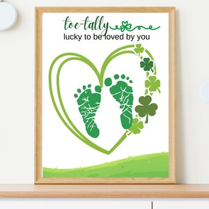May include: A framed print with a white background features green baby footprints inside a green heart. The words "toe-tally lucky to be loved by you" are written above the heart. Green shamrocks decorate the right side of the heart.