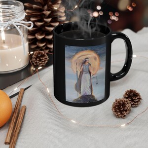 May include: A black ceramic mug featuring a painting of a robed figure holding a sword and scales, set against a golden sun. The mug has a curved handle and is filled with a dark liquid, possibly coffee or tea.
