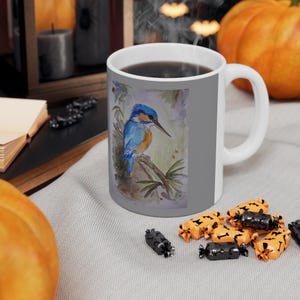 Könnte beinhalten: Eine weiße Keramiktasse mit grauem Hintergrund und einer Aquarell-Illustration eines blauen und orangefarbenen Eisvogels, der auf einem Ast sitzt. Die Tasse hat einen schwarzen Henkel und ist mit Kaffee gefüllt.