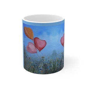 Könnte beinhalten: Ein weißer Keramik-Kaffeebecher mit einem Gemälde von herzförmigen Ballons, die in einem blauen Himmel über einem Feld mit Wildblumen schweben. Die Ballons sind in Rosa- und Orangetönen gehalten.