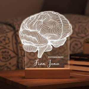 May include: A lighted brain-shaped lamp with the name "Fina Jean" and the word "Psychologist" etched on the clear acrylic panel. The lamp sits on a wooden base and emits a warm glow, suitable for a desk or bedside table.