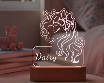 May include: A glowing unicorn night light with the name "Dairy" etched on the front. The unicorn design is white and sits atop a rectangular wooden base. The light is on a nightstand next to a bed.