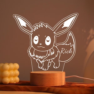 Może przedstawiać: Biała lampka nocna z postacią Eevee i imieniem "Rick" wygrawerowanym z boku. Światło stoi na drewnianej podstawie i jest podłączone do przewodu zasilającego. Tło jest ciepło brązowe.