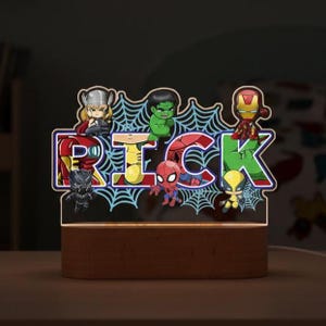 Op de afbeelding: Een gepersonaliseerd nachtlampje met de naam "RICK" in een kleurrijk, verlicht ontwerp. Het licht bevat superhelden zoals Thor, Hulk, Iron Man en Spider-Man, tegen een webachtergrond, op een houten voet.