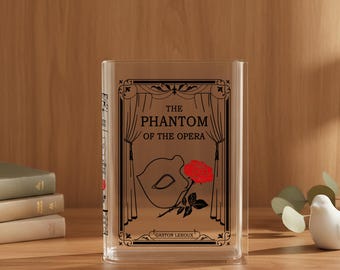 May include: Clear, rectangular book-shaped container with the text "THE PHANTOM OF THE OPERA" and a graphic of a mask and rose. The container is made of clear material and has a black border. The author's name, Gaston Leroux, is at the bottom.