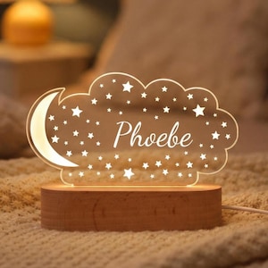 May include: A night light shaped like a cloud with a crescent moon and stars. The name "Phoebe" is written in a script font. The light sits on a wooden base and is plugged in.