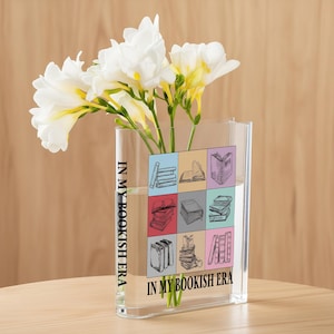 May include: A clear rectangular vase with white and yellow flowers. The vase has a colorful book-themed design with the text "IN MY BOOKISH ERA". The vase is filled with water and the flowers are fresh.