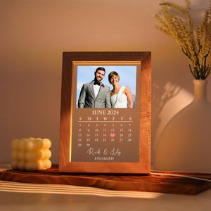 May include: Wooden framed photo with a picture of a couple, a calendar for June 2024, and the text "Rick & Lily ENGAGED". The calendar highlights June 19th. The frame is illuminated with a warm glow.