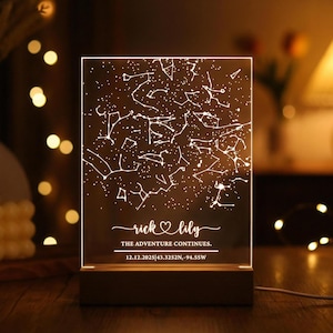 Puede incluir: Una luz nocturna personalizada con un diseño de mapa estelar. El panel acrílico transparente muestra un patrón de constelaciones con el texto "rick & lily" y "THE ADVENTURE CONTINUES." La base es de madera. También se muestra la fecha y las coordenadas.