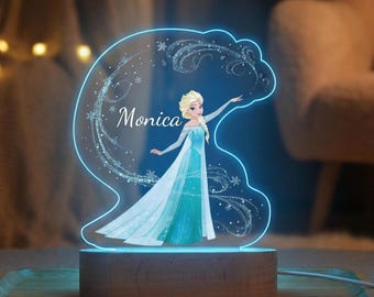 Op de afbeelding: Een gepersonaliseerde LED-nachtlamp met een cartoonafbeelding van een prinses in een blauw-witte jurk. De naam "Monica" is in cursief geschreven. Het licht staat op een houten voet en straalt een zachte blauwe gloed uit.