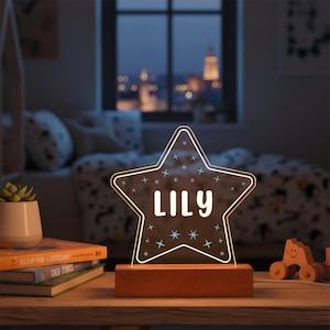 May include: A star-shaped night light with the name "LILY" in white lettering. The light has a wooden base and is decorated with small blue stars. The light is on a wooden table with books and a small plant.