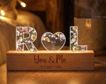 May include: A wooden night light featuring the letters "R" and "L" and a heart-shaped design. The letters and heart are adorned with floral patterns. The base of the light is engraved with the words "You & Me" and "WE GOT THIS".