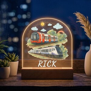 May include: An illuminated night light with a wooden base, showcasing two train illustrations. The top train is red and white, labeled "Piccadilly", and the bottom train is white and purple, labeled "Elizabeth". The name "RICK" is displayed below.