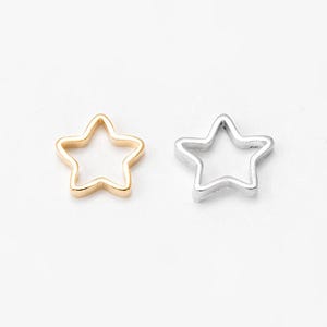 May include: Two star-shaped charms. One is gold-colored, the other is silver-colored. Both charms have a hollow, open design. The charms are placed on a white background. These charms could be used for jewelry making or other craft projects.