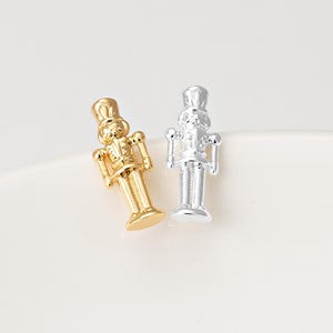 May include: Two small nutcracker figurines, one finished in a polished gold tone and the other in a bright silver tone. Each figurine features a tall hat, a uniform jacket with detailed buttons, and a cylindrical base, standing upright.