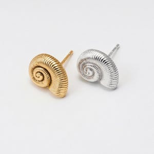 May include: Two seashell-shaped stud earrings. One earring is gold-colored, and the other is silver-colored. The earrings have a spiral design and are set against a white background. The earrings are small and delicate.