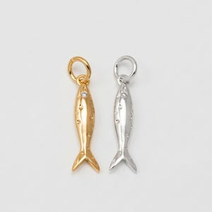Könnte beinhalten: Zwei fischförmige Anhänger, einer goldfarben und einer silberfarben, vor weißem Hintergrund. Jeder Anhänger hat eine kleine Schlaufe oben und einen kleinen Edelstein. Der goldfarbene Anhänger hat ein warmes, glänzendes Finish.