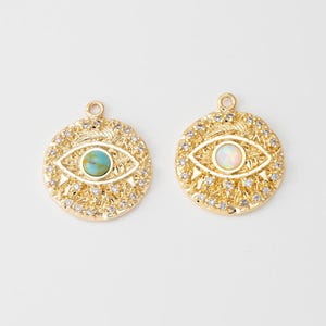 May include: Two circular gold-tone pendants featuring an eye motif. Each pendant has a central cabochon stone, one turquoise and one iridescent white, surrounded by small clear crystals set in a textured, sunburst-like gold metal frame.