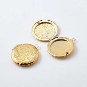 Op de afbeelding: Een gouden hanger met een bloemmotief aan de voorkant en een effen achterkant. De hanger is open en de achterkant is zichtbaar.