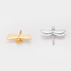 Op de afbeelding: Twee libellenbedels. Eén is goudkleurig, met gedetailleerde vleugels en lichaam. De andere is zilverkleurig, ook met gedetailleerde vleugels en een lus om op te hangen. Beide tegen een witte achtergrond.