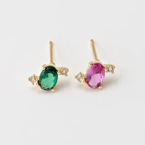 May include: A pair of stud earrings featuring an oval-cut green gemstone and an oval-cut pink gemstone. Each earring has a gold-colored post and is accented with small, clear gemstones. The earrings are set against a white background.