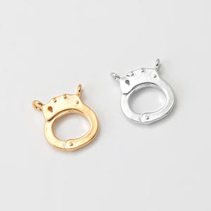 Puede incluir: Dos cierres metálicos, uno dorado y otro plateado, se muestran sobre un fondo blanco. Los cierres tienen forma de esposas en miniatura, con una abertura circular y pequeños bucles para la fijación. Estos cierres son adecuados para la fabricación de joyas.