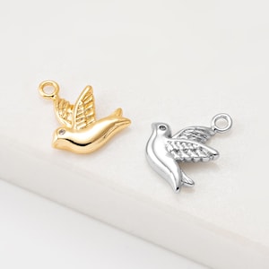 Pode incluir: Dois pingentes de metal em forma de pomba, um dourado e outro prateado, sobre um fundo branco. Cada pingente apresenta um pássaro com as asas abertas e uma pequena argola na parte superior para prender a joias ou outros trabalhos manuais.