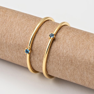 Peut inclure: Deux bracelets dorés, chacun orné d'une petite pierre précieuse bleue ronde. Les bracelets sont incurvés et reposent sur une surface cylindrique marron. Le fond est blanc uni.