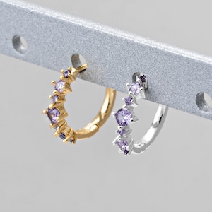Op de afbeelding: Twee oorringen, één goudkleurig en één zilverkleurig, elk versierd met kleine, paarse edelstenen. De oorbellen worden getoond op een zilveren staaf met cirkelvormige uitsparingen.
