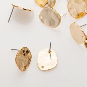 Puede incluir: Una colección de pendientes de botón dorados con textura martillada. Los pendientes tienen un cierre de poste y una forma ovalada ligeramente irregular. El metal tiene un acabado brillante y reflectante. Los pendientes están esparcidos sobre una superficie blanca.