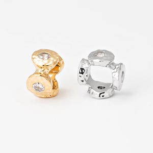 May include: Two decorative jewelry charms. One is gold-toned with a textured surface and a clear gem. The other is silver-toned with a textured surface and a clear gem. Both charms have a unique, open design.