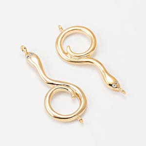 Puede incluir: Dos colgantes dorados con forma de serpiente, con un cuerpo enrollado y un pequeño acento de cristal en la cabeza. Los colgantes están diseñados con un lazo en la parte superior e inferior para sujetarlos a las joyas. El fondo es blanco.