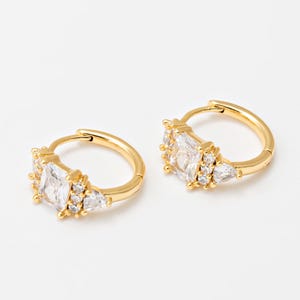 May include: A pair of gold-colored hoop earrings. Each earring features a central, square-cut clear stone, flanked by smaller, clear, triangular and round stones. The earrings are set against a white background.