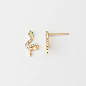 Puede incluir: Un par de pendientes de botón dorados con forma de serpiente. La cabeza de la serpiente está adornada con una pequeña gema verde, y el cuerpo está incrustado con pequeños cristales transparentes. Sobre fondo blanco.